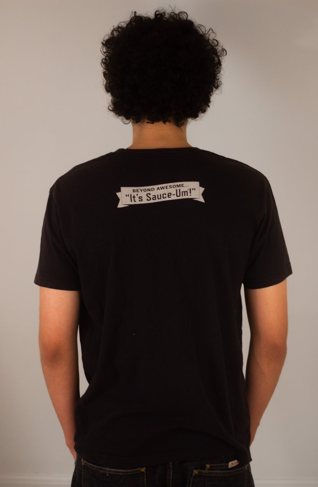 Person wearing a black t-shirt with text, "Beyond awesome...it's sauce-um! "on the back against a plain background