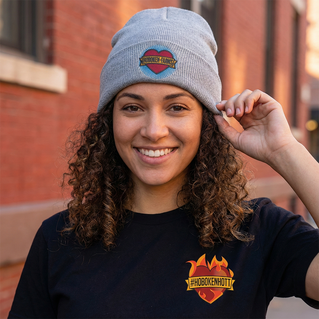 Person wearing a gray beanie with a Hoboken Farms logo and black t-shirt with a Hoboken Farms logo, standing in front of a brick building.